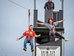 A young woman and young man are harnessed in a zip line and speed toward the camera.